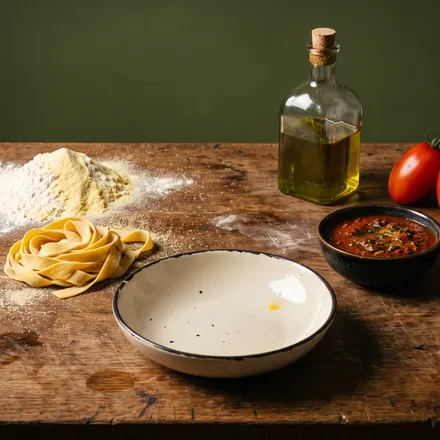 Fresh pasta nests, tomatoes, flour and olive oil laid out on a wooden bench